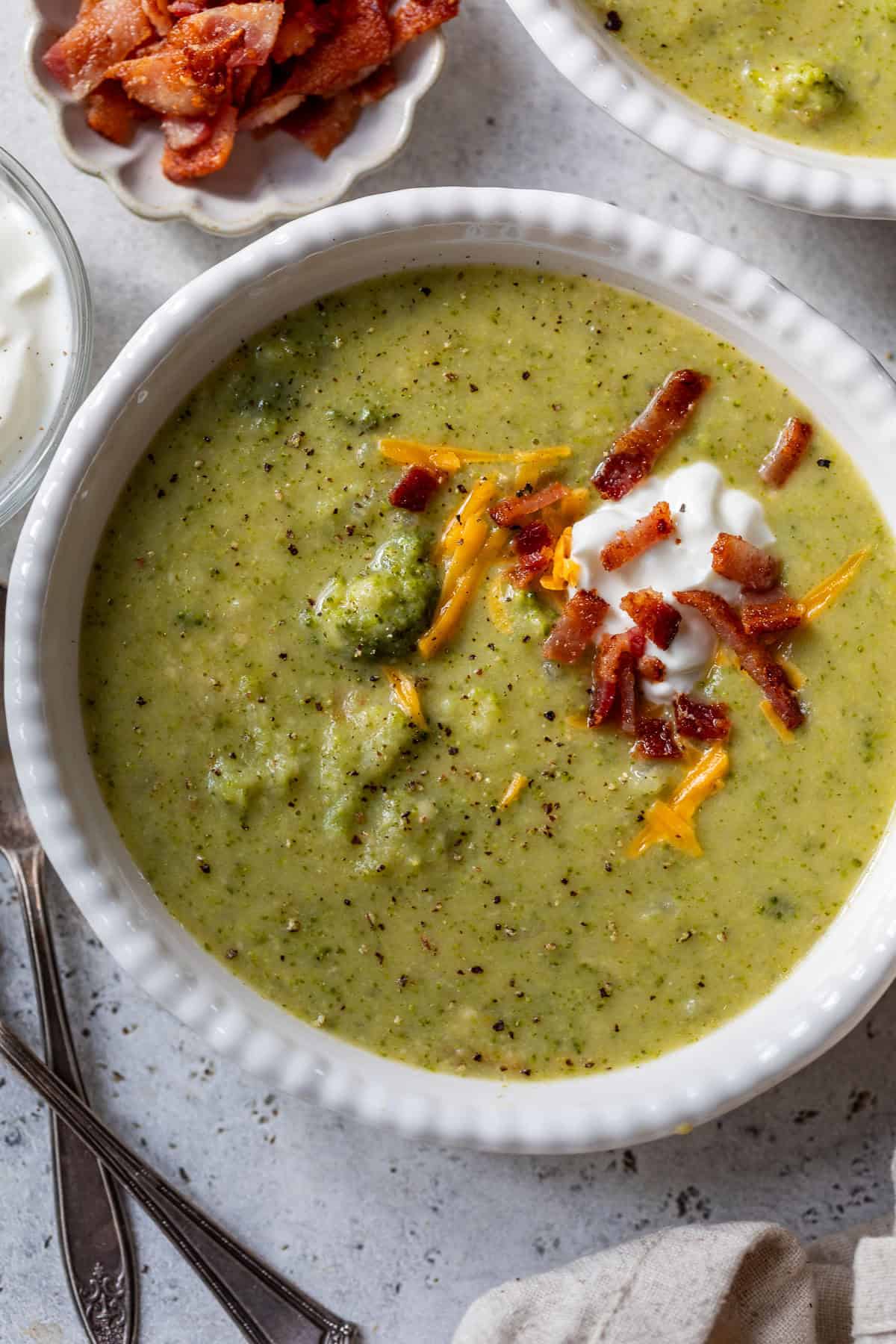 Bowl of broccoli potato soup topped with sour cream, shredded cheese, and crumbled bacon, with a spoon beside it.