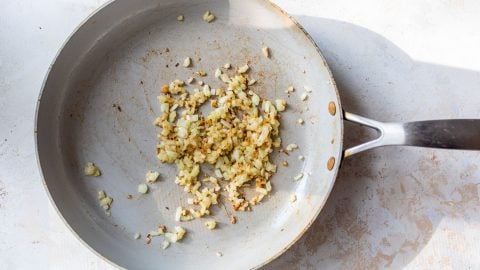 Chopped onions being sautéed and lightly browned in a metal frying pan on a light surface.