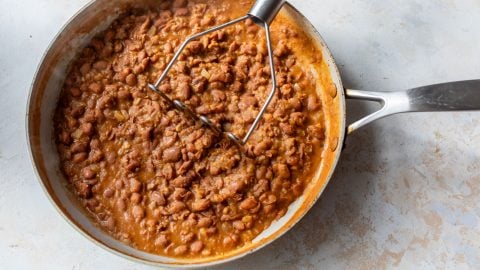 A skillet of refried beans being mashed with a metal potato masher on a light-colored surface.
