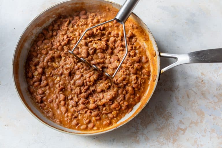 A skillet of refried beans being mashed with a metal potato masher on a light-colored surface.