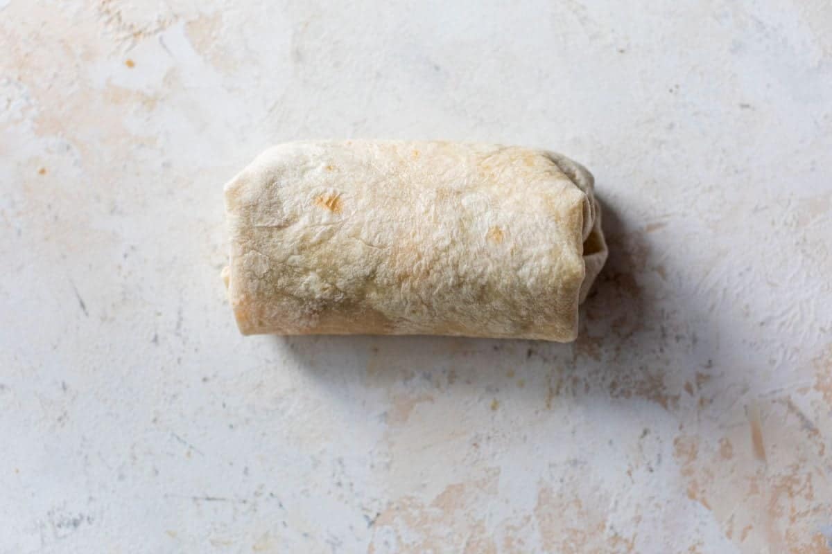 A single wrapped burrito sits on a textured, light-colored surface.