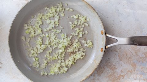 Diced onions cooking in a large silver skillet on a light-colored countertop.