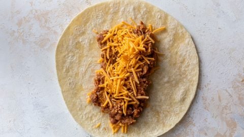 Flour tortilla topped with refried bean mixture and shredded cheddar cheese on a light surface.