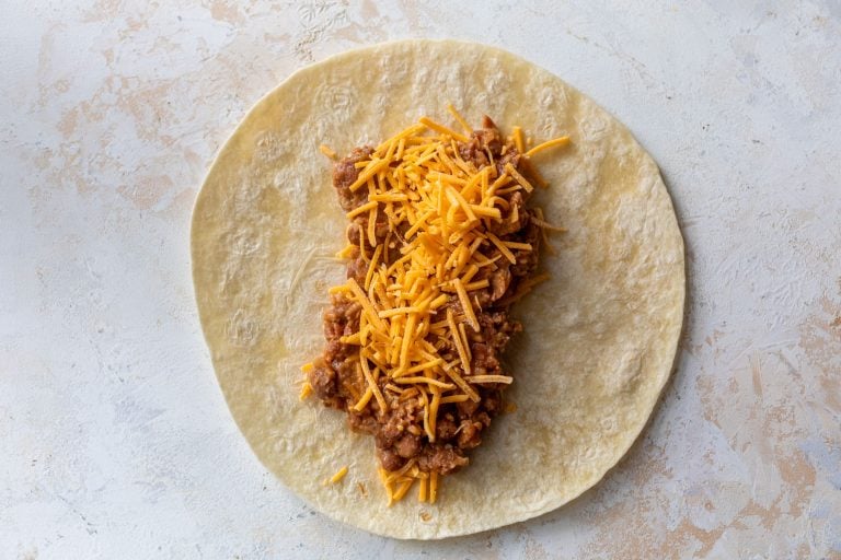Flour tortilla topped with refried bean mixture and shredded cheddar cheese on a light surface.
