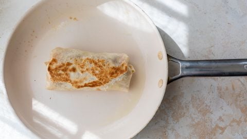 A browned burrito cooking in a white frying pan, sunlight casting striped shadows on the surface.