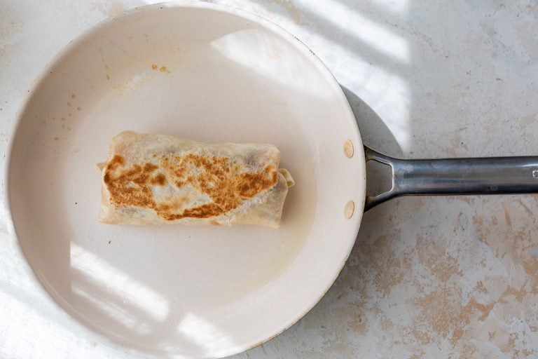 A browned burrito cooking in a white frying pan, sunlight casting striped shadows on the surface.