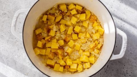 Diced potatoes and onions cooking in a white pot on a gray countertop, with sunlight casting shadows.