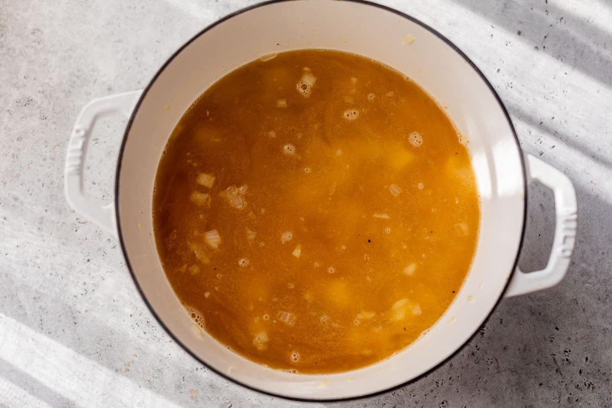 A white pot filled with broth and chopped onions sits on a light gray countertop in sunlight.