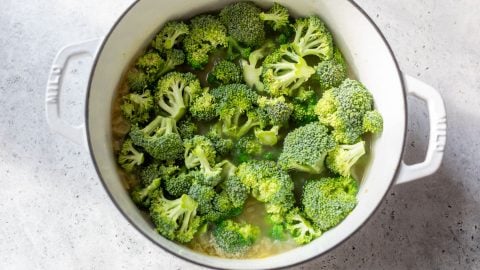 Broccoli florets cooking in a white pot with handles on a light grey countertop.