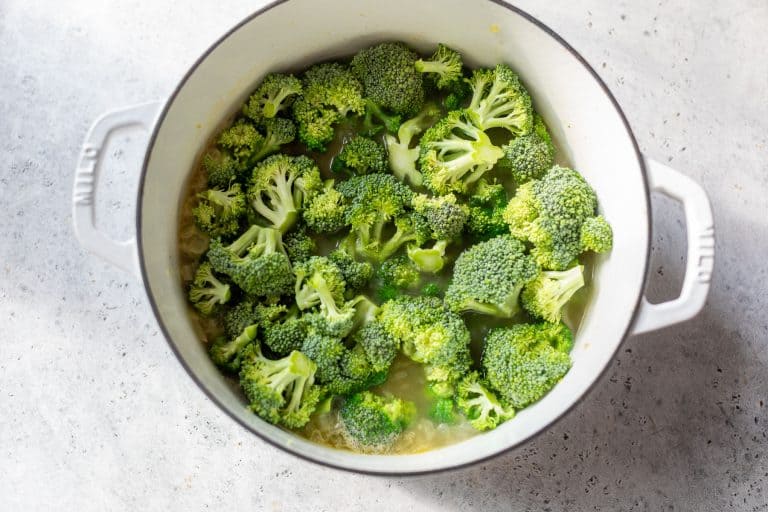 Broccoli florets cooking in a white pot with handles on a light grey countertop.