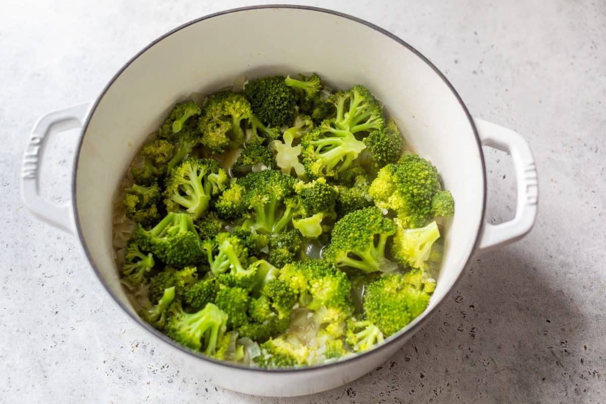 Broccoli florets cooking in a white pot with chopped onions on a light gray surface.