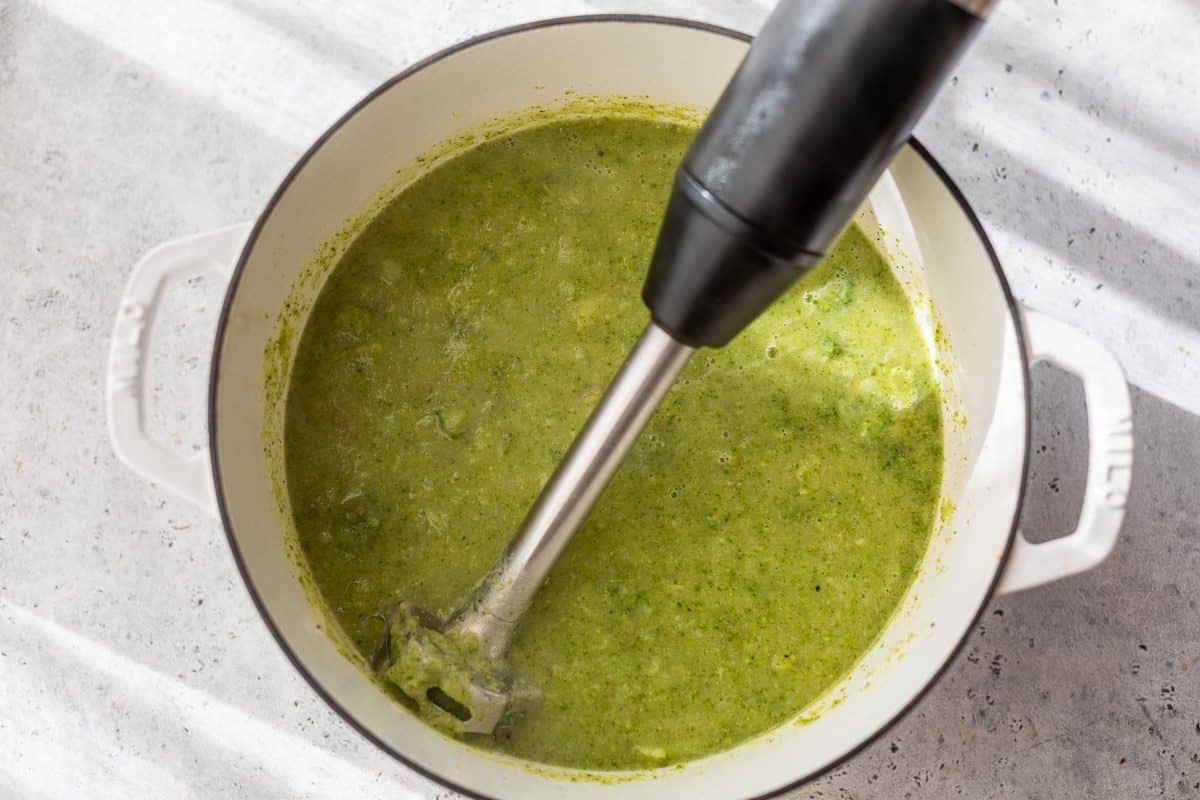 An immersion blender pureeing broccoli potato soup in a white pot on a light countertop.