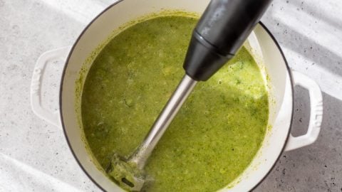 An immersion blender pureeing broccoli potato soup in a white pot on a light countertop.