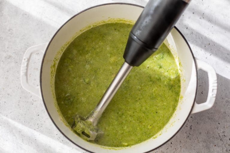 An immersion blender pureeing broccoli potato soup in a white pot on a light countertop.