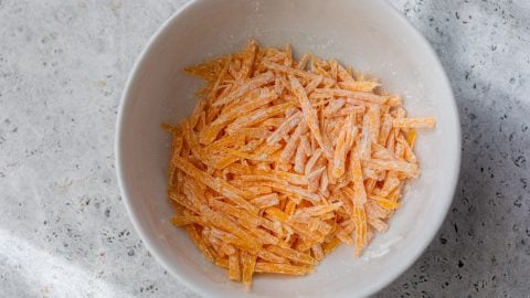 Shredded cheddar coated in cornstarch in a white bowl on a light textured surface.