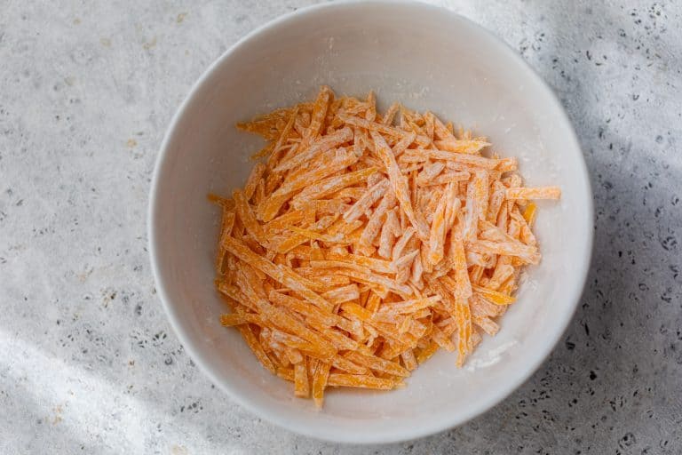 Shredded cheddar coated in cornstarch in a white bowl on a light textured surface.