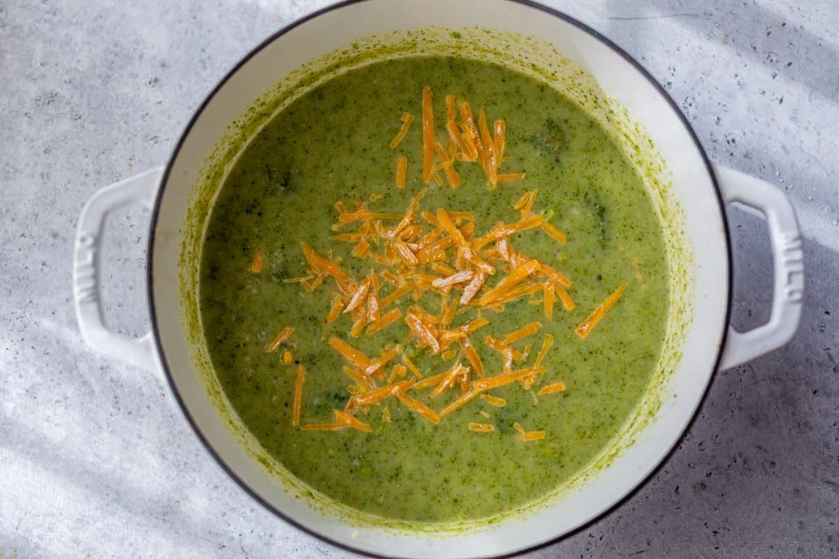 Broccoli potato soup in a white pot topped with shredded cheddar cheese, sitting on a gray surface.