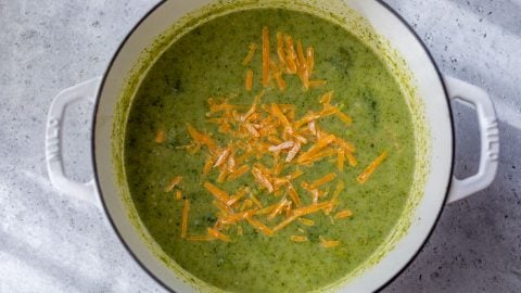 Broccoli potato soup in a white pot topped with shredded cheddar cheese, sitting on a gray surface.