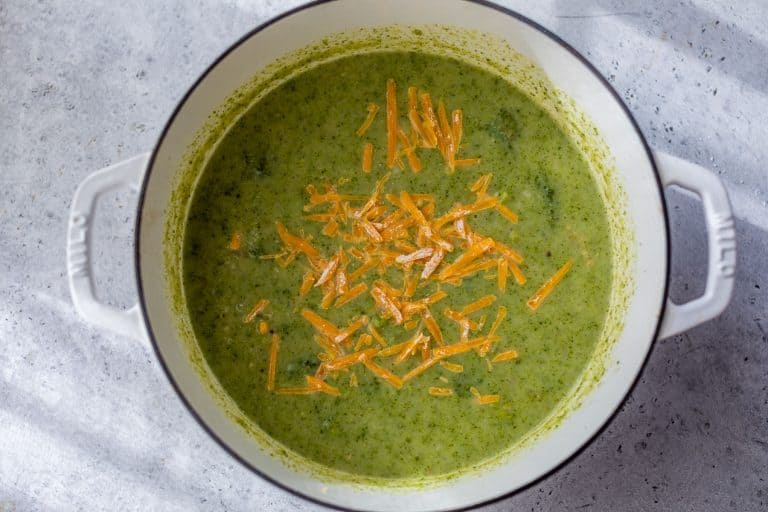 Broccoli potato soup in a white pot topped with shredded cheddar cheese, sitting on a gray surface.