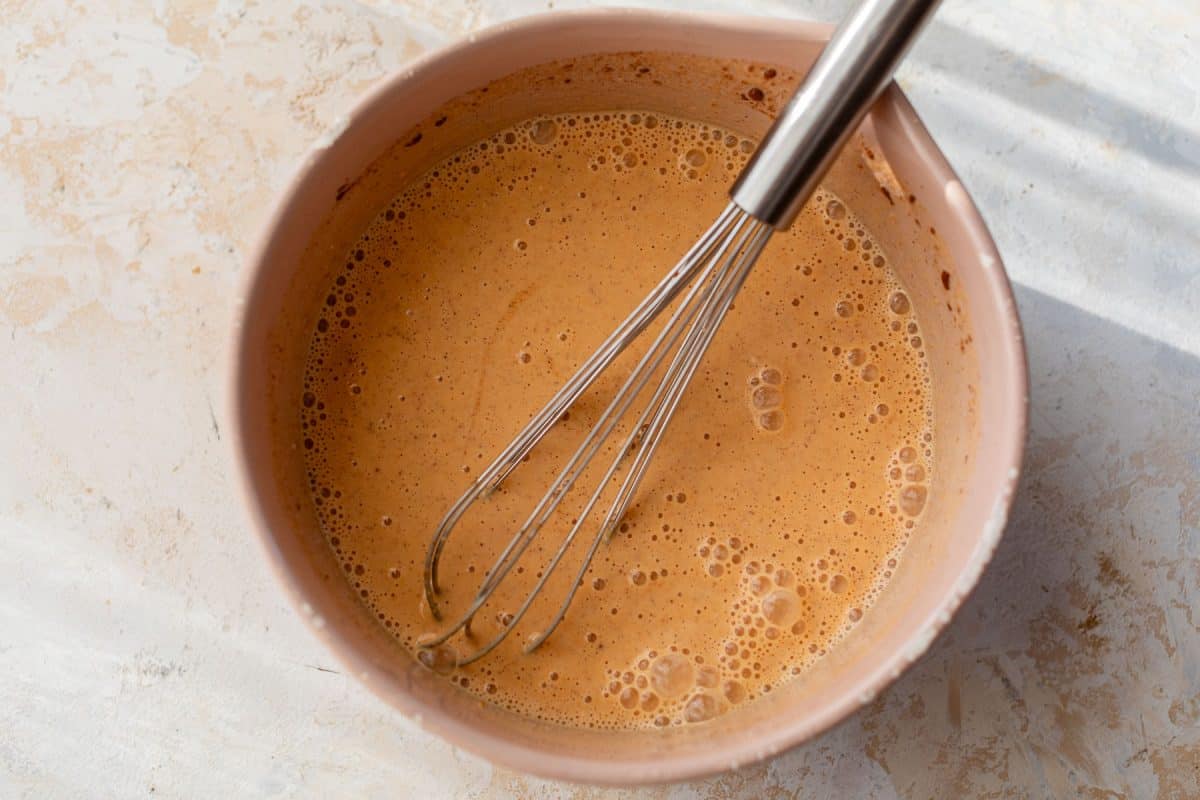A metal whisk in a bowl of light brown pumpkin mixture on a textured, light-colored surface.