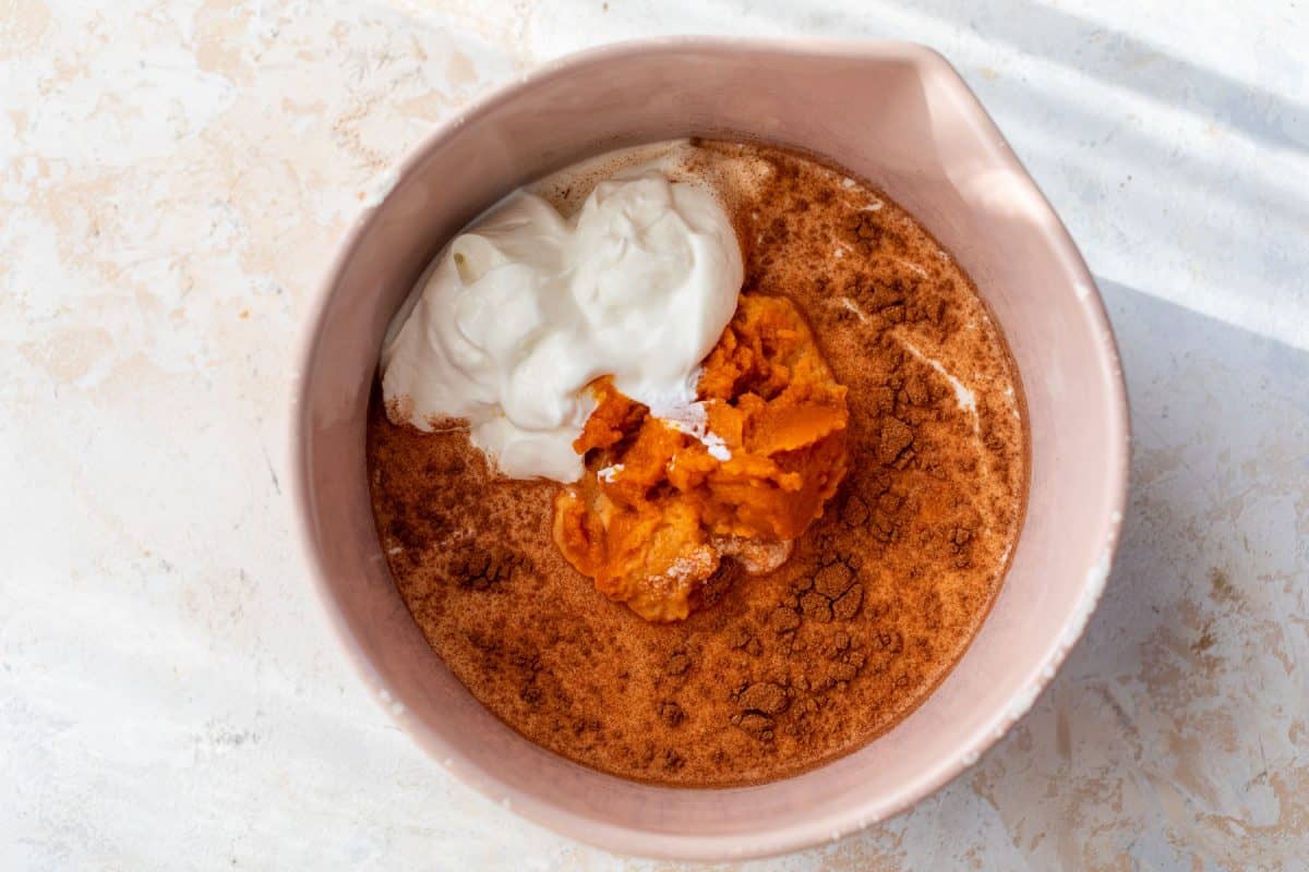 A pink bowl with pumpkin puree, yogurt, and cinnamon on a light surface, ready to be mixed.
