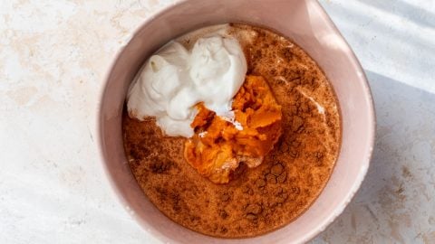 A pink bowl with pumpkin puree, yogurt, and cinnamon on a light surface, ready to be mixed.