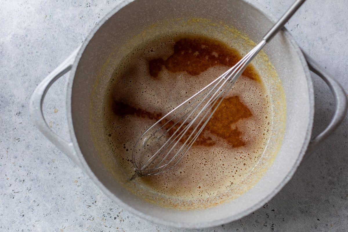 A metal whisk rests in a gray pot containing a light brown, foamy liquid on a gray surface.
