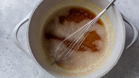 A metal whisk rests in a gray pot containing a light brown, foamy liquid on a gray surface.