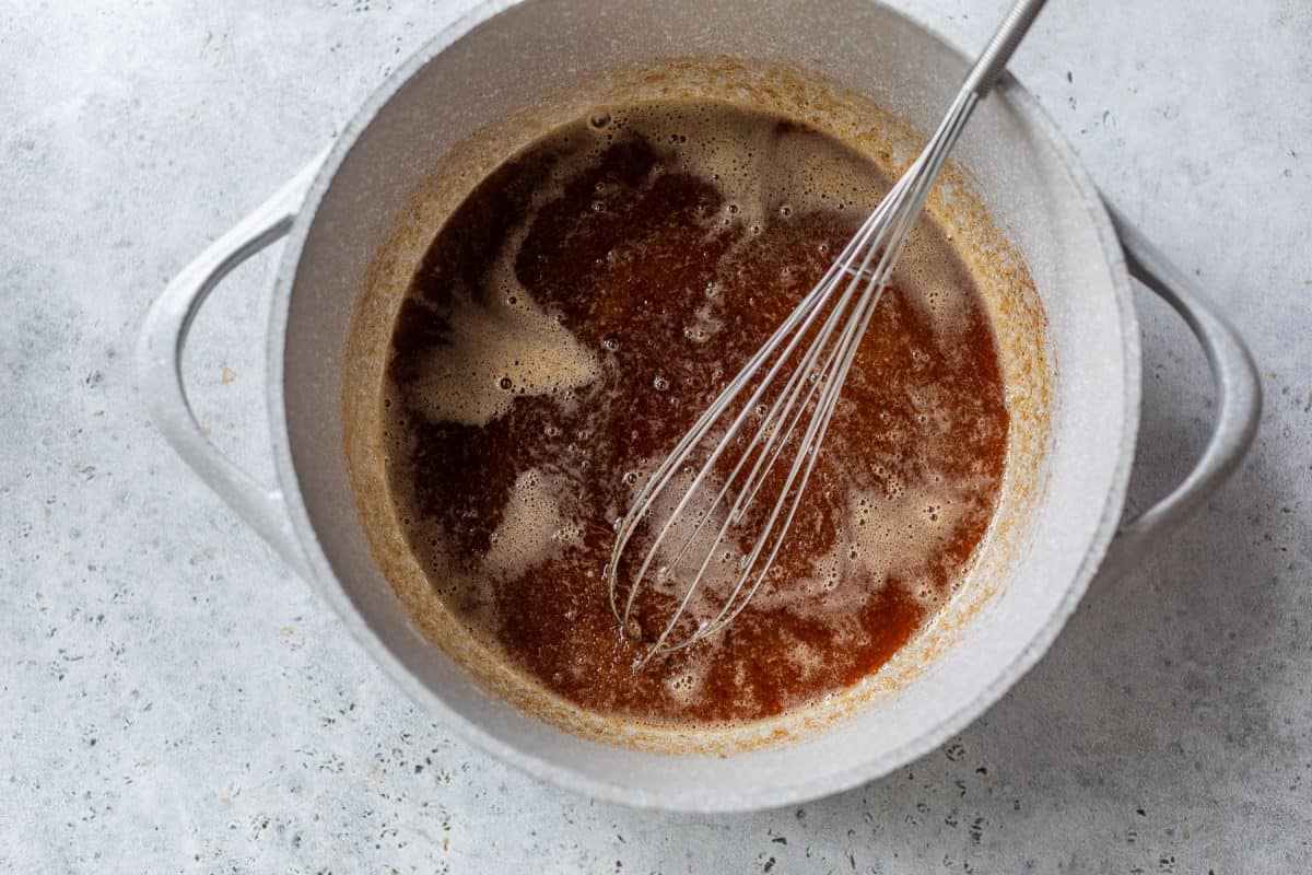 A metal whisk in a pot of melted browned butter on a light gray countertop.