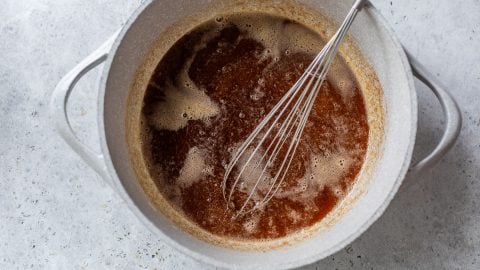 A metal whisk in a pot of melted browned butter on a light gray countertop.