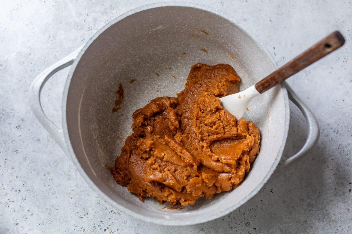 Thick pumpkin cookie dough mixture in a white pot with a spatula on a light gray textured surface.