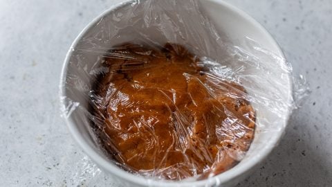 Brown pumpkin spice cookie dough in a white bowl covered with plastic wrap, resting on a light gray surface.