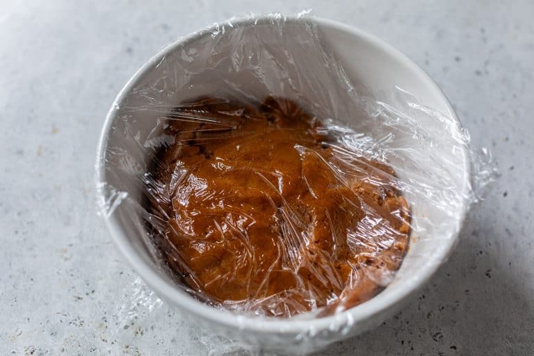 Brown pumpkin spice cookie dough in a white bowl covered with plastic wrap, resting on a light gray surface.