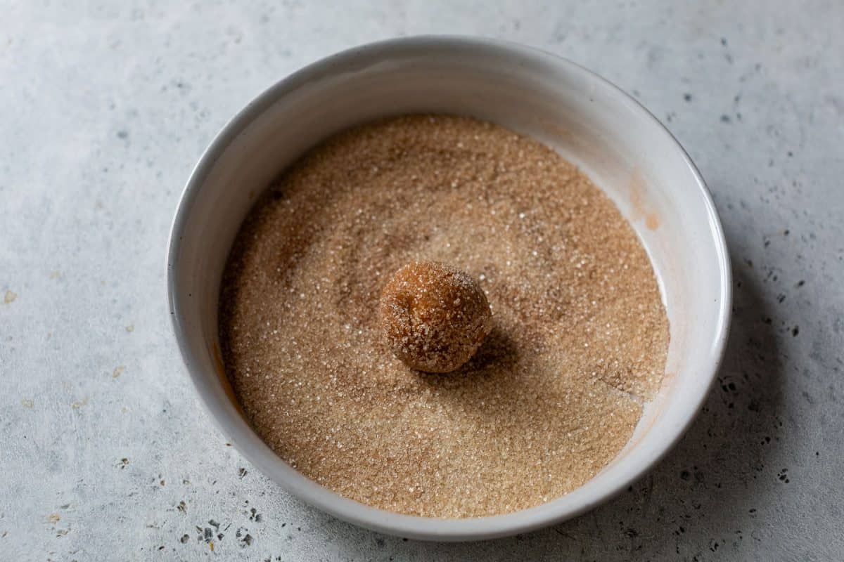 A ball of cookie dough is coated in cinnamon sugar in a white bowl on a gray surface.