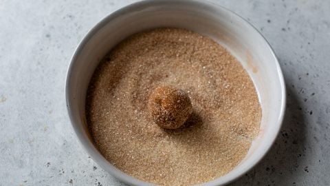 A ball of cookie dough is coated in cinnamon sugar in a white bowl on a gray surface.