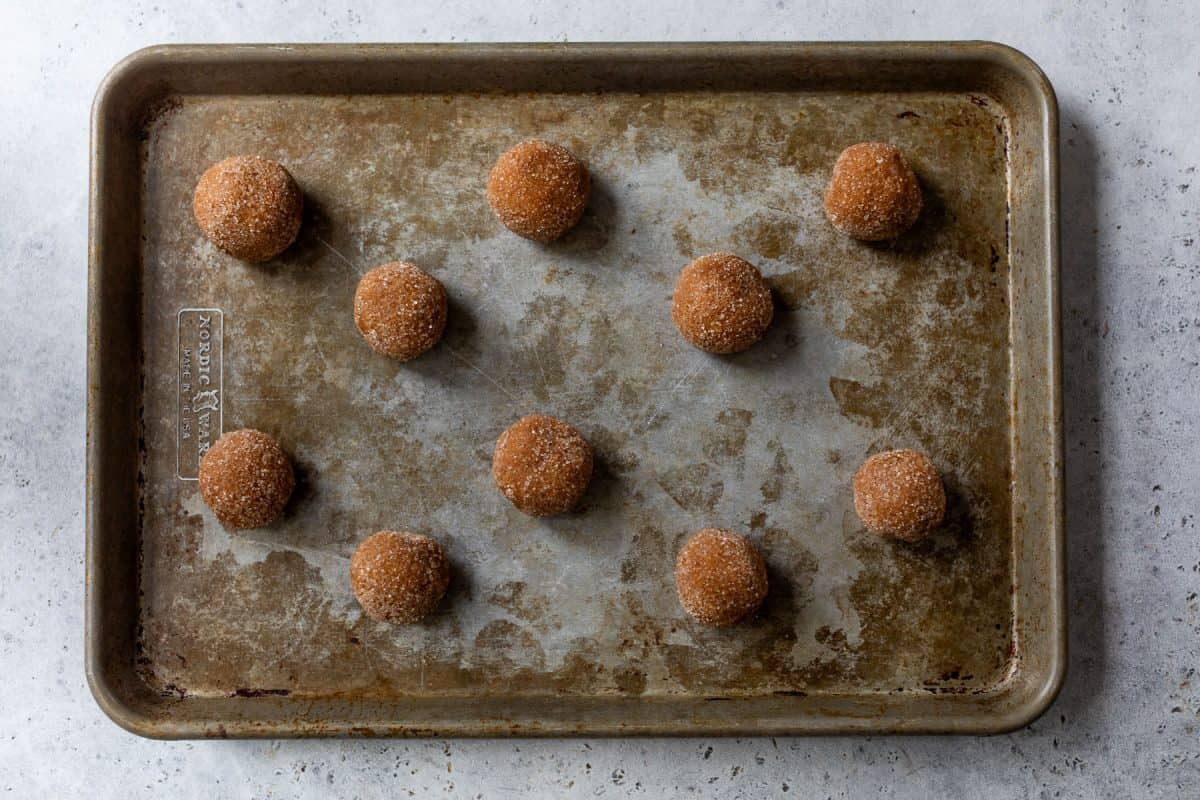 Twelve round pumpkin spice cookie dough balls coated in cinnamon sugar on a worn metal baking sheet.