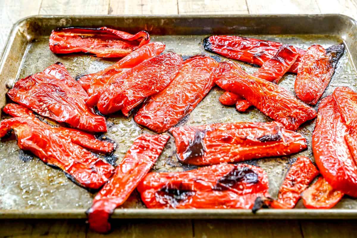 Red bell pepper slices roasted on a metal baking sheet, with charred edges and shiny, slightly wrinkled skin.