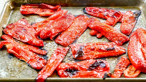 Red bell pepper slices roasted on a metal baking sheet, with charred edges and shiny, slightly wrinkled skin.