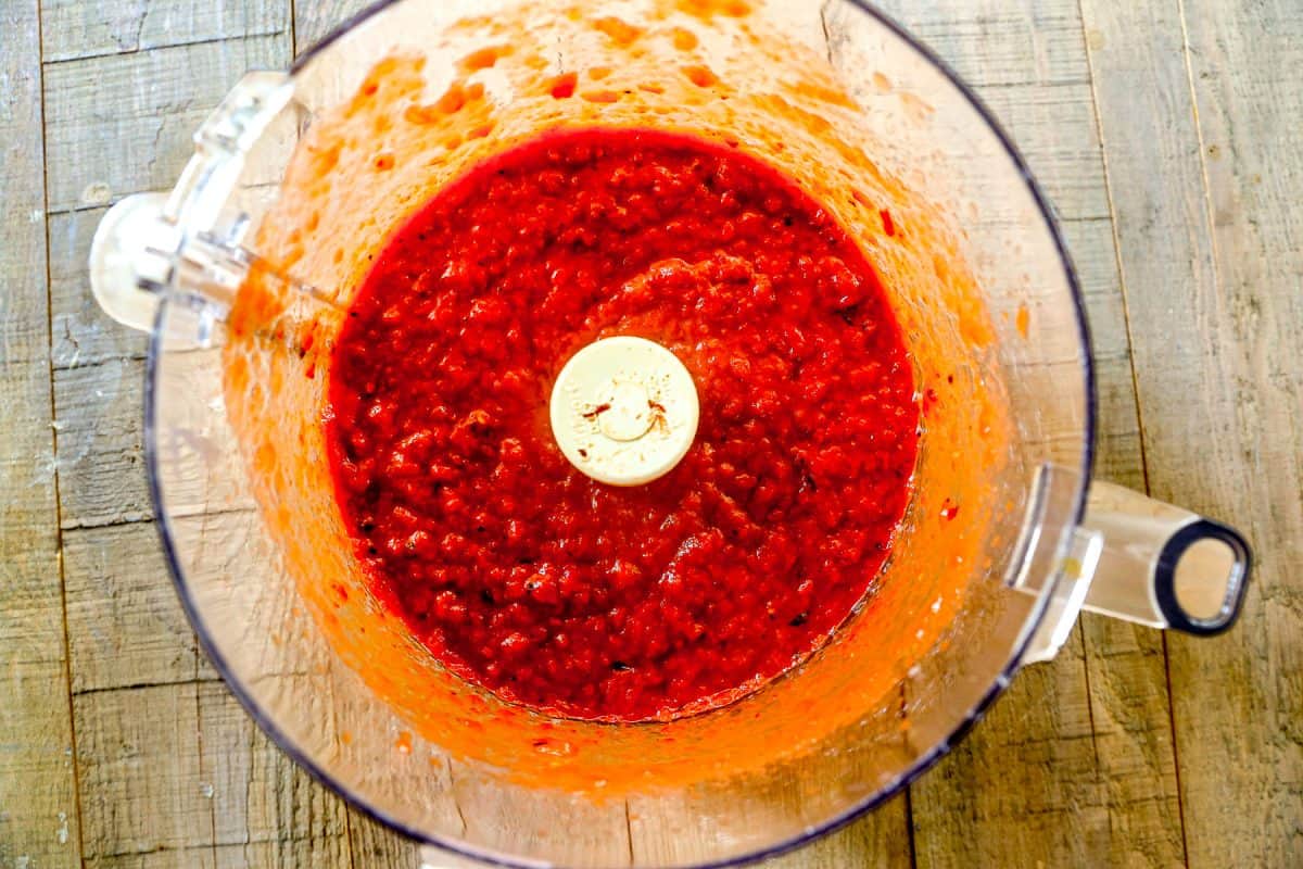 Bright red pepper and tomato mixture blended in a food processor bowl, viewed from above on a wooden surface.