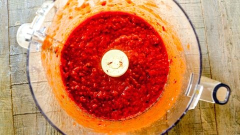 Bright red pepper and tomato mixture blended in a food processor bowl, viewed from above on a wooden surface.