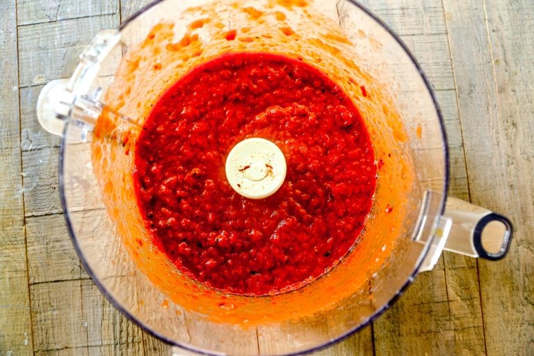 Bright red pepper and tomato mixture blended in a food processor bowl, viewed from above on a wooden surface.