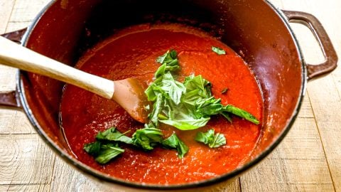 Roasted red pepper soup in a pot with fresh basil leaves and a wooden spoon on a wooden table.