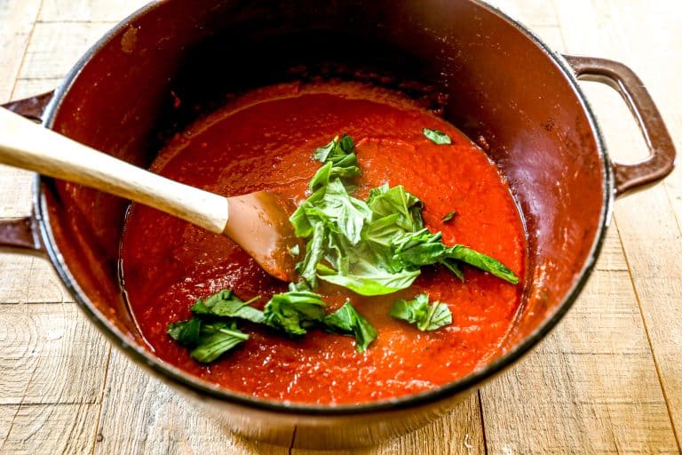 Roasted red pepper soup in a pot with fresh basil leaves and a wooden spoon on a wooden table.