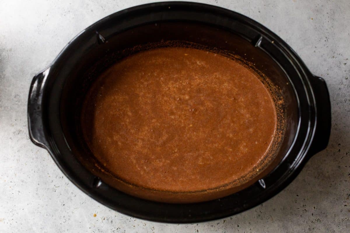 Ingredients for pumpkin oatmeal mixed in a black slow cooker on a gray textured surface.