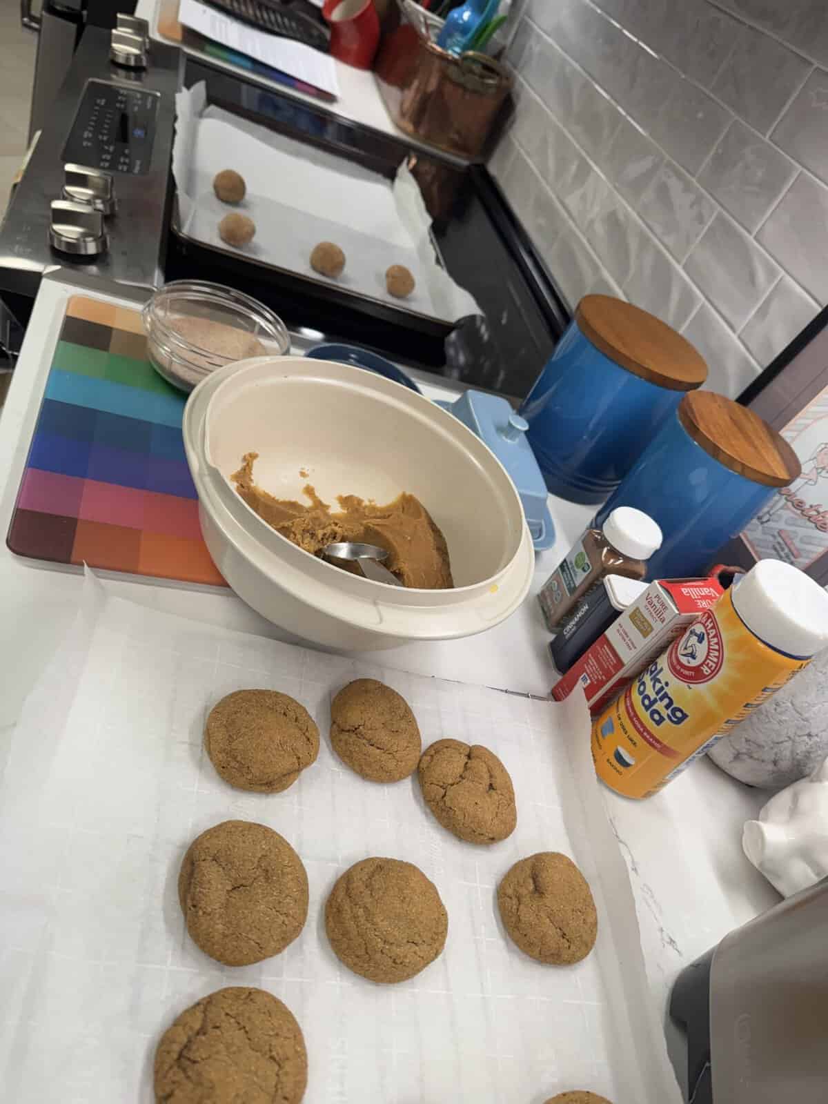 Baking scene with cookies on trays, a bowl of dough, measuring cups, and kitchen tools on a countertop.