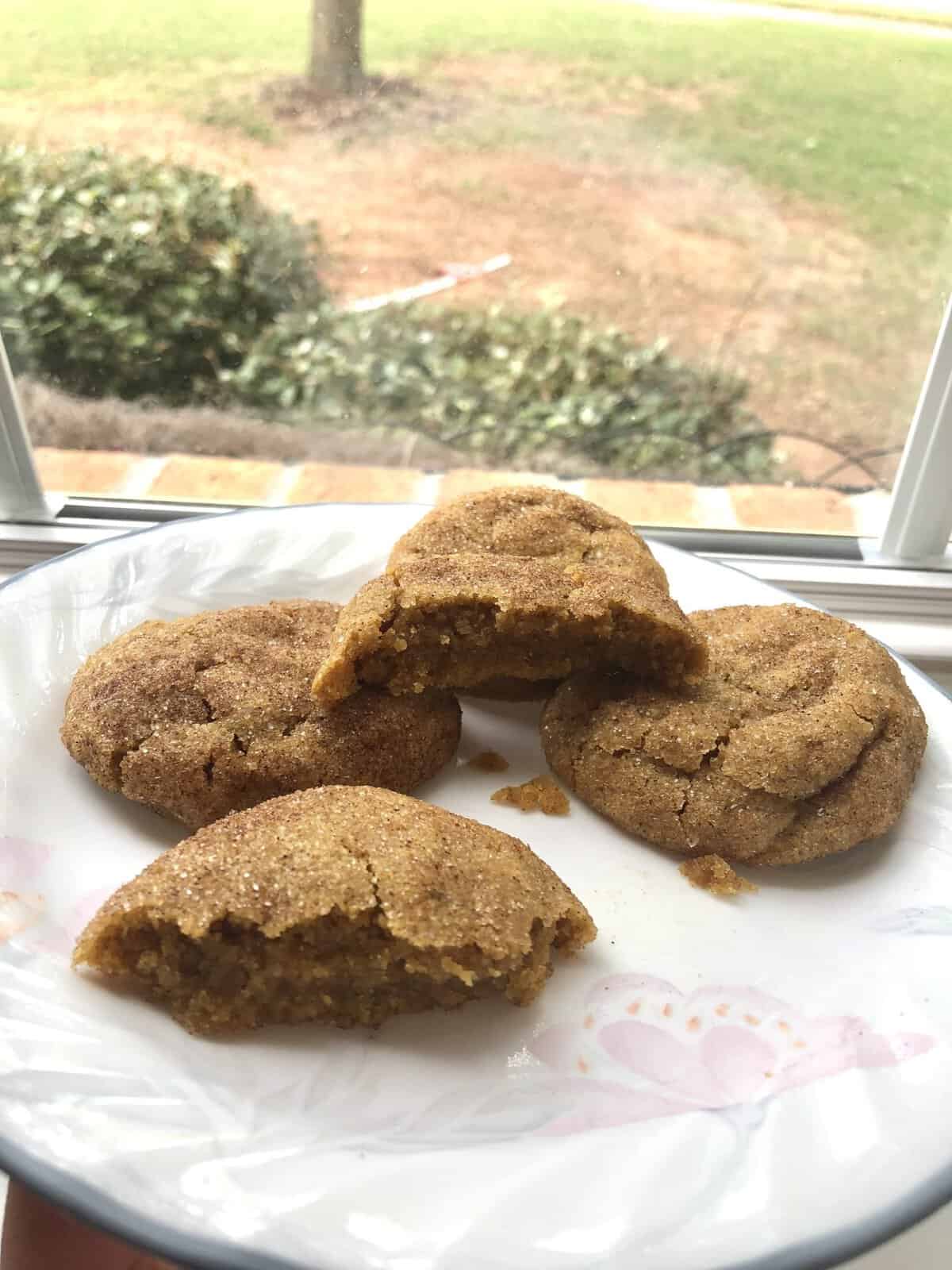 Four brown sugar cookies, one broken in half, on a white plate by a window with a grassy yard outside.