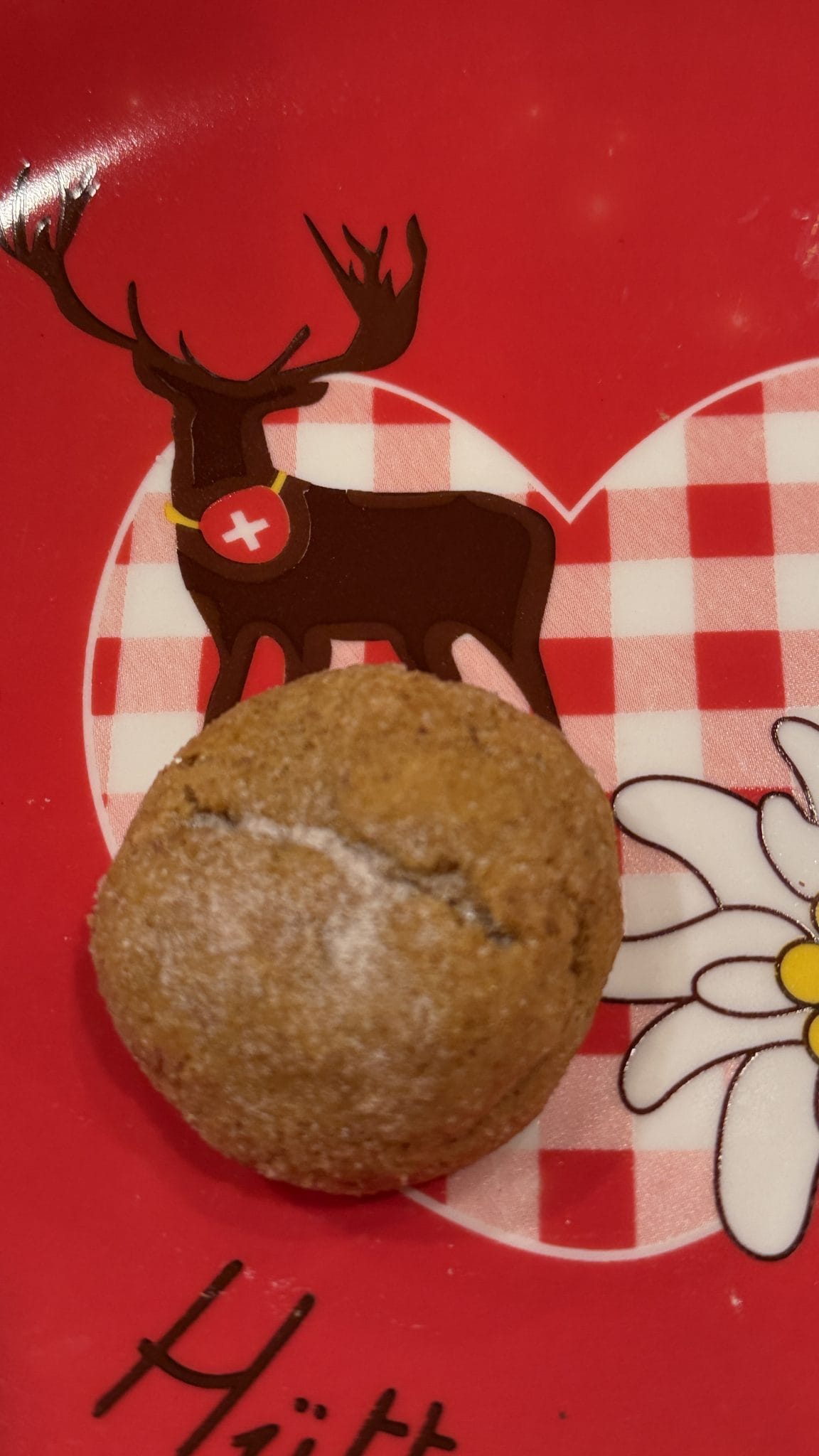 A brown cookie sits on a red plate with a deer, Swiss cross, heart, and white flower design.
