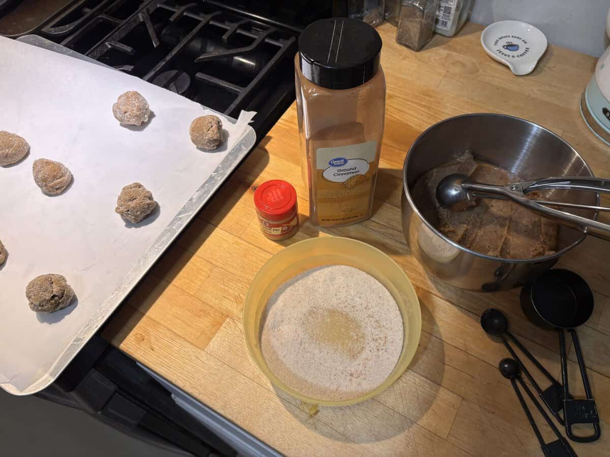 Cookie dough balls on a baking tray, cinnamon, nutmeg, and a bowl of cinnamon sugar on a kitchen counter.