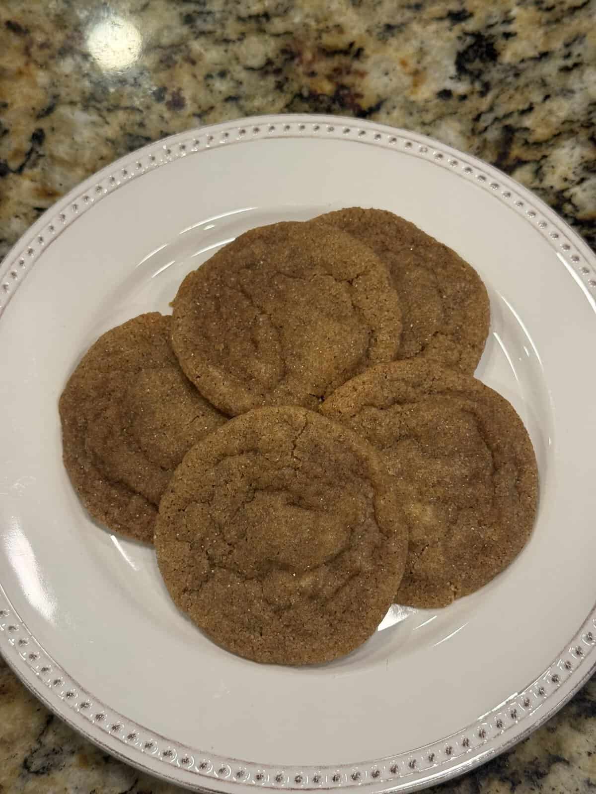 Five brown cookies are arranged on a white plate with a decorative rim, placed on a granite countertop.