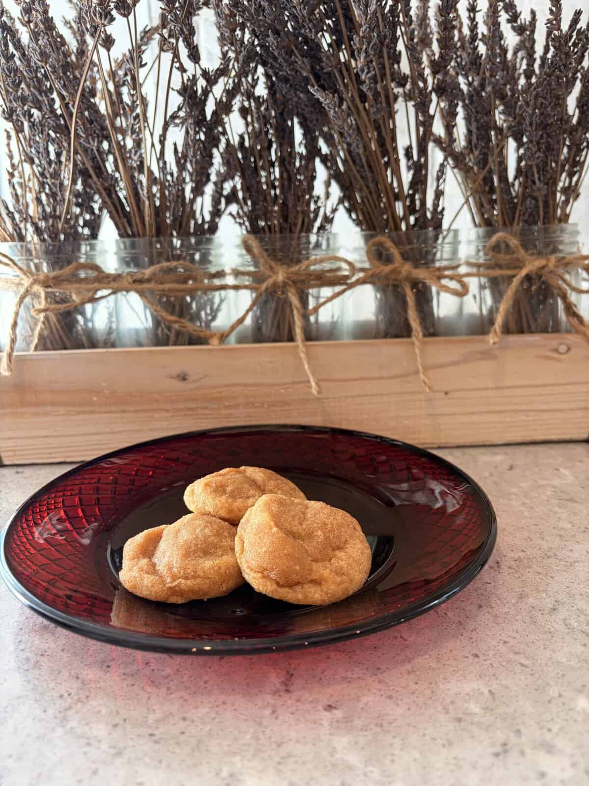Three cookies on a red glass plate sit on a countertop with dried lavender in jars tied with twine in the background.